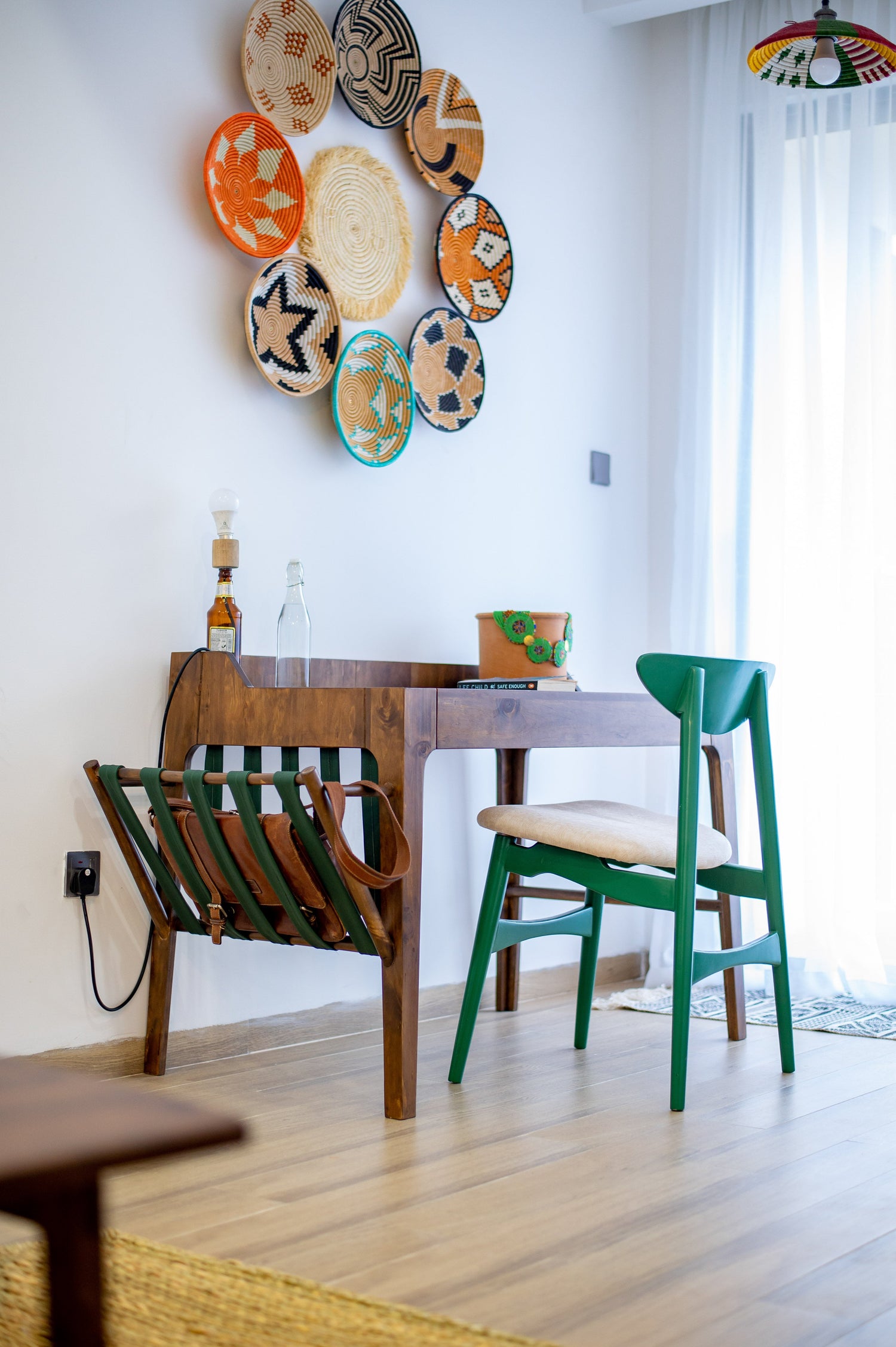 The Manda Desk — a handcrafted solid wood desk with a warm walnut finish, green accent chair, and built-in leather magazine holder in a bright modern room.